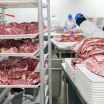 Trabalhadores manuseiam cortes de carne em bandejas em uma unidade industrial de processamento de alimentos.